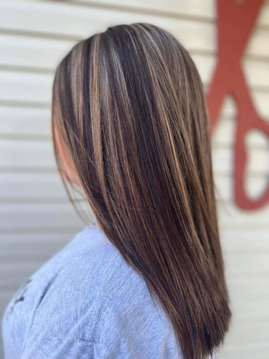Long dark hair worn straight with warm caramel highlights, photographed outside the salon beside one of the oversized scissors mounted on the wall.